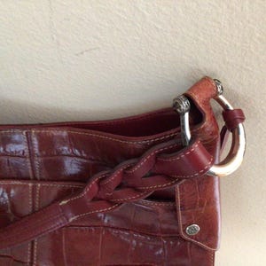 May include: A close-up of a reddish-brown leather handbag with a crocodile skin texture. The bag features a braided strap and silver-toned hardware. The bag has a small, round silver emblem.
