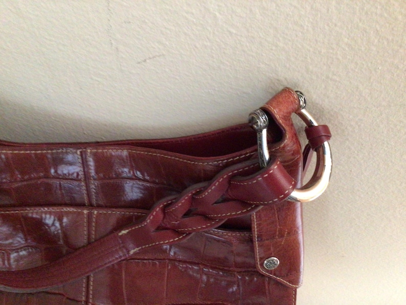 May include: A close-up of a reddish-brown leather handbag with a crocodile skin texture. The bag features a braided strap and silver-toned hardware. The bag has a small, round silver emblem.