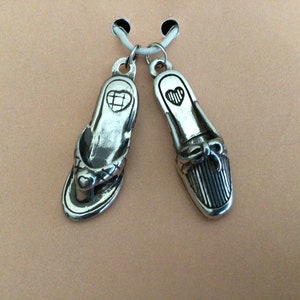 Pode incluir: Dois pingentes de prata em forma de sapatos. Um &eacute; uma sand&aacute;lia com um design de cora&ccedil;&atilde;o e o outro &eacute; um sapato fechado com um la&ccedil;o e listras.