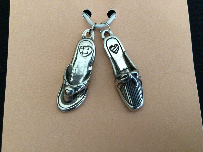 Pode incluir: Dois pingentes de prata em forma de sapatos. Um &eacute; uma sand&aacute;lia com um design de cora&ccedil;&atilde;o e o outro &eacute; um sapato fechado com um la&ccedil;o e listras.