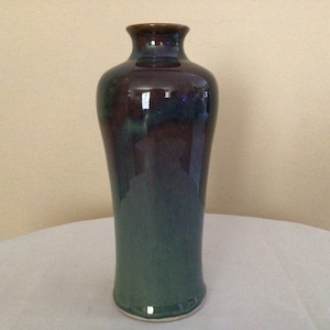 May include: A tall, slender ceramic vase with a dark purple and green glaze. The vase has a rounded bottom and a narrow opening at the top.