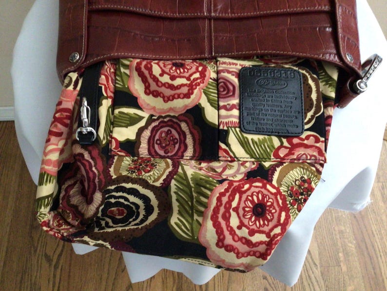 May include: A brown leather handbag with a floral patterned interior. The bag features a textured exterior and a silver-colored clasp. The interior fabric displays a pattern of red, pink, and cream-colored flowers on a black background.