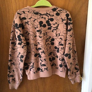 Op de afbeelding: Een roze sweatshirt met ronde hals en lange mouwen, met een herhaald patroon van zwarte en witte Mickey Mouse-ontwerpen. Het sweatshirt hangt aan een groene hanger voor een houten deur.