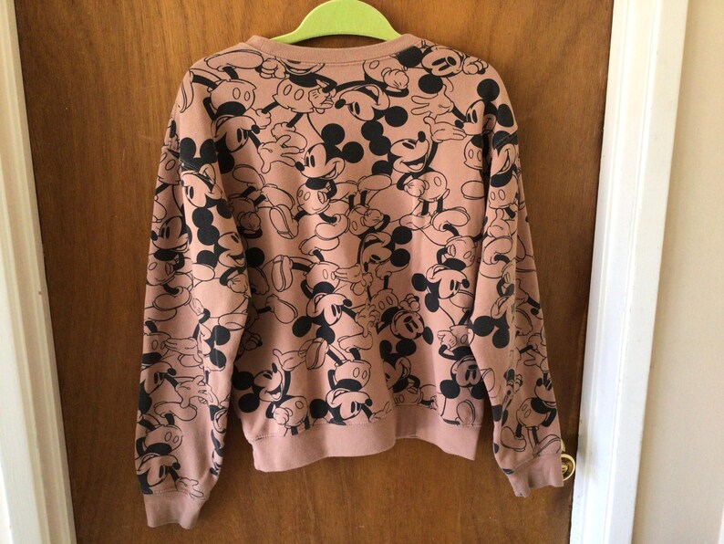 Op de afbeelding: Een roze sweatshirt met ronde hals en lange mouwen, met een herhaald patroon van zwarte en witte Mickey Mouse-ontwerpen. Het sweatshirt hangt aan een groene hanger voor een houten deur.