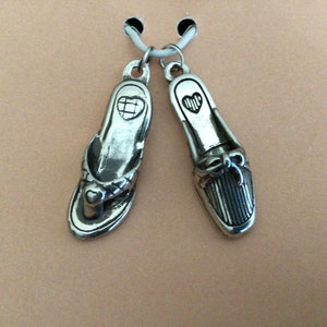 Pode incluir: Dois pingentes de prata em forma de sapatos. Um &eacute; uma sand&aacute;lia de dedo com um detalhe de cora&ccedil;&atilde;o e o outro &eacute; um mocassim com um la&ccedil;o.