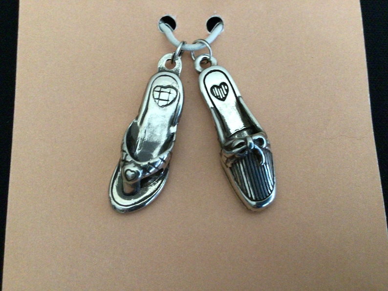 Pode incluir: Dois pingentes de prata em forma de sapatos. Um &eacute; uma sand&aacute;lia de dedo com um detalhe de cora&ccedil;&atilde;o e o outro &eacute; um mocassim com um la&ccedil;o.