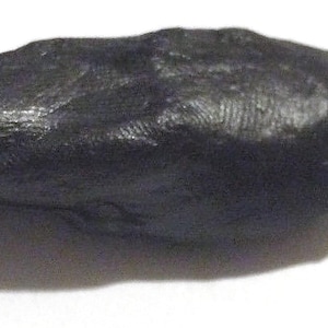 May include: A dark charcoal gray, elongated, oval-shaped stone with a textured, matte surface. The object features subtle, fine, linear striations across its top and sides, giving it a natural, weathered appearance with rounded, smooth edges.