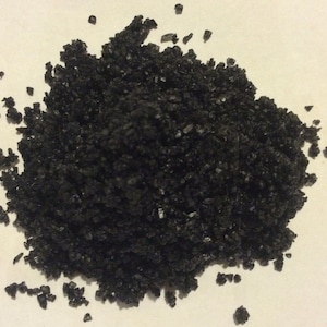 May include: A close-up of black Hawaiian sea salt. The salt is coarse and has a dark, almost black color. It is a natural product and is often used in cooking and as a finishing salt.