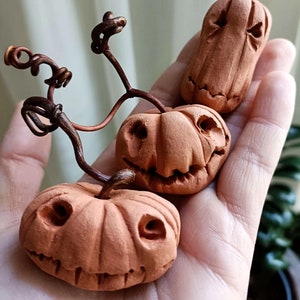 May include: Three small, terracotta-coloured clay pumpkins with carved, expressive faces and dark brown, twisted wire stems. The pumpkins feature textured surfaces, deep vertical grooves, and hollowed-out eyes and mouths, arranged in a cluster.