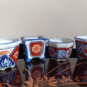 May include: Four small ceramic plant pots with a white, red, and blue floral pattern. The pots are in different shapes, including round and square, and are displayed on a dark, reflective surface.