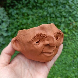 May include: A terracotta-coloured ceramic planter shaped like a human face with a smiling expression, prominent nose, and textured eyebrows. The vessel features a hollow top opening and a rough, earthy finish, showcasing detailed facial features.