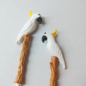 May include: Two decorative stirrers or picks, each featuring a white cockatoo with a yellow crest and a black beak, perched atop a textured brown branch. The birds are sculpted with detailed feathers and are positioned vertically on the stems.