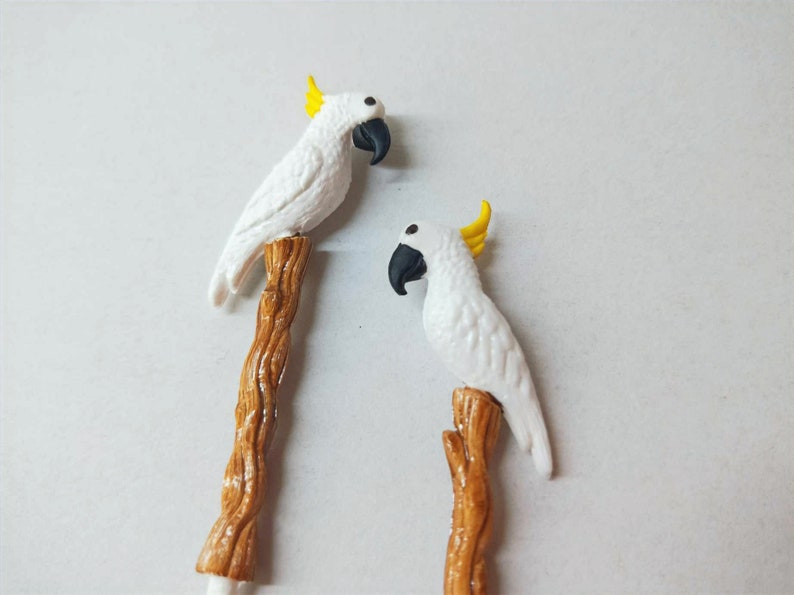May include: Two decorative stirrers or picks, each featuring a white cockatoo with a yellow crest and a black beak, perched atop a textured brown branch. The birds are sculpted with detailed feathers and are positioned vertically on the stems.