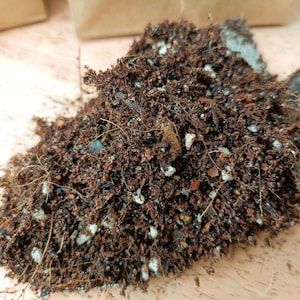 Puede incluir: Primer plano de un mont&oacute;n de tierra para macetas de color marr&oacute;n oscuro con peque&ntilde;as part&iacute;culas blancas y rojas mezcladas. La tierra est&aacute; suelta y desmenuzable, con algunas finas ra&iacute;ces marrones visibles.