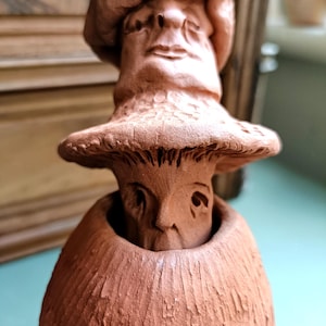 May include: A terracotta mushroom-shaped sculpture with a face carved into the cap and stem. The sculpture has a textured surface and a whimsical, artistic design. The mushroom figure is a unique decorative item.