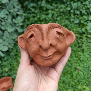 May include: A terracotta-colored, face-shaped planter with a smiling expression, large ears, and detailed facial features. The planter is held in a hand against a backdrop of green foliage. Two additional planters are visible in the background.