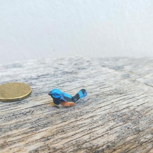 May include: A small, hand-painted blue bird figurine with black and orange details, displayed on a textured wooden surface. A gold coin is placed nearby for size comparison. The bird is intricately crafted.