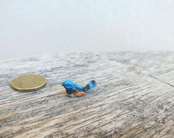 May include: A miniature blue bird figurine with black and orange accents, perched on a weathered wooden surface. A gold coin is next to the bird for scale. The bird has a detailed, hand-painted appearance.