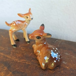 May include: Two ceramic deer figurines. One deer is standing with brown and white spots, the other is sitting with brown and white spots.