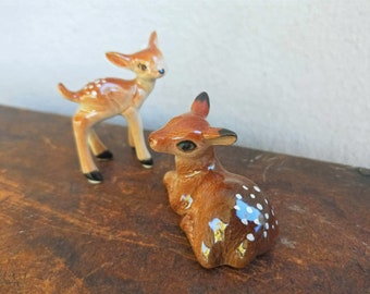 May include: Two ceramic deer figurines. One deer is standing with brown and white spots, the other is sitting with brown and white spots.