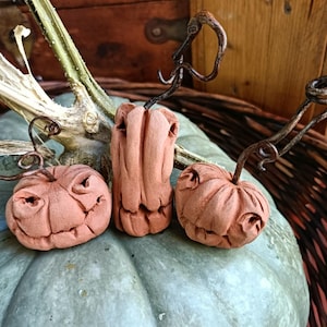 May include: Three small, hand-sculpted terracotta-coloured clay figures with carved facial features and dark, twisted metal wire stems. Two figures are round and pumpkin-shaped, while the central figure is tall and cylindrical in shape.