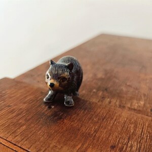 Może przedstawiać: Mała, ciemna figurka ceramiczna przedstawiająca wombat z błyszczącym wykończeniem. Zwierzę jest przedstawione w pozycji stojącej, posiada spiczaste uszy, jasny pysk, ciemny nos oraz widoczną teksturę futra na zaokrąglonym tułowiu figurki.