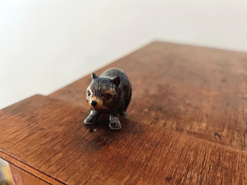 Może przedstawiać: Mała, ciemna figurka ceramiczna przedstawiająca wombat z błyszczącym wykończeniem. Zwierzę jest przedstawione w pozycji stojącej, posiada spiczaste uszy, jasny pysk, ciemny nos oraz widoczną teksturę futra na zaokrąglonym tułowiu figurki.