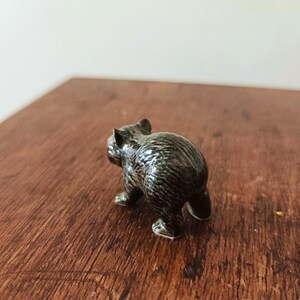 Może przedstawiać: Mała, ciemnobrązowa ceramiczna figurka niedźwiedzia widziana od tyłu. Niedźwiedź ma błyszczące, teksturowane wykończenie imitujące futro, z widocznymi wyrzeźbionymi uszami i czterema łapami stojącymi stabilnie na płaskiej powierzchni.