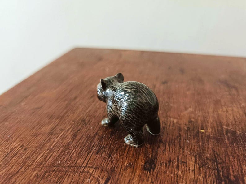 Może przedstawiać: Mała, ciemnobrązowa ceramiczna figurka niedźwiedzia widziana od tyłu. Niedźwiedź ma błyszczące, teksturowane wykończenie imitujące futro, z widocznymi wyrzeźbionymi uszami i czterema łapami stojącymi stabilnie na płaskiej powierzchni.