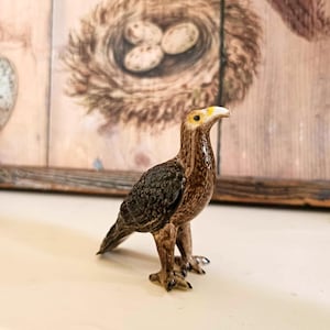 May include: A ceramic figurine of a brown and black hawk with yellow eyes. The hawk is standing on a white surface.