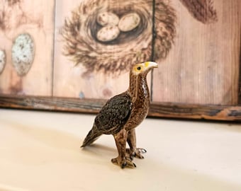 May include: A ceramic figurine of a brown and black hawk with yellow eyes. The hawk is standing on a white surface.
