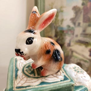 May include: A ceramic figurine of a brown and white spotted rabbit with pink ears. The rabbit is sitting on a green and white ceramic base with intricate designs.