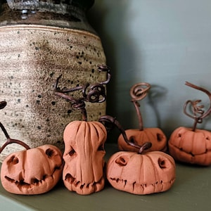 May include: Four small, hand-sculpted terracotta pumpkins with carved facial features and dark, twisted wire stems. Each pumpkin displays a unique expression, ranging from wide grins to jagged, toothy smiles, arranged in a group on a flat surface.