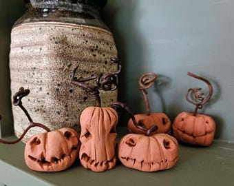 May include: Four small, hand-sculpted terracotta pumpkins with carved facial features and dark, twisted wire stems. Each pumpkin displays a unique expression, ranging from wide grins to jagged, toothy smiles, arranged in a group on a flat surface.