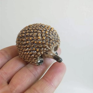 May include: A detailed, miniature ceramic echidna figurine. The figurine features a textured, brown body with small, yellow-orange accents. It has a long, dark snout and small, dark eyes. The echidna is held in a hand against a plain white background.