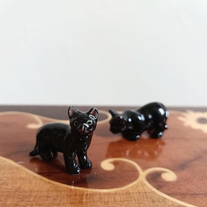 May include: Two small, glossy black cat figurines. One cat is standing, the other is in a seated position. The figurines are on a wooden surface with decorative inlaid patterns. The cats have detailed features.