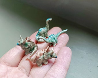 Puede incluir: Cuatro figuritas de dinosaurio de cerámica en miniatura. Dos son dinosaurios rayados de verde y azul, uno es un dinosaurio rayado de marrón y blanco, y otro es un triceratops marrón y blanco.