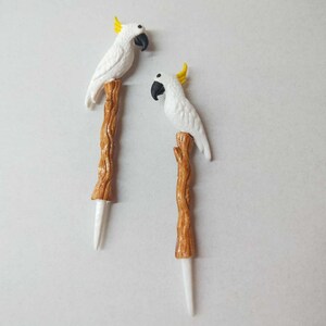 May include: Two decorative picks, each featuring a white cockatoo with a yellow crest and a black beak, perched atop a textured brown branch-shaped stem that tapers into a smooth white point at the bottom end of the object.