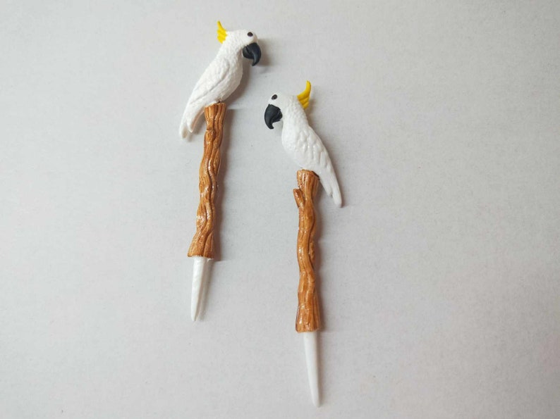 May include: Two decorative picks, each featuring a white cockatoo with a yellow crest and a black beak, perched atop a textured brown branch-shaped stem that tapers into a smooth white point at the bottom end of the object.