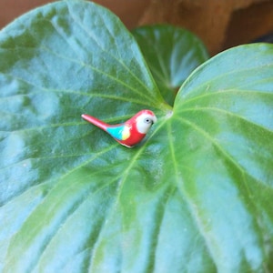 May include: A small, red, blue and yellow ceramic bird figurine perched on a green leaf.