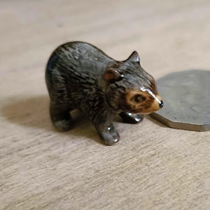 May include: A small ceramic figurine of a grey and brown possum with black eyes and a white nose. The figurine is standing on a wooden surface.