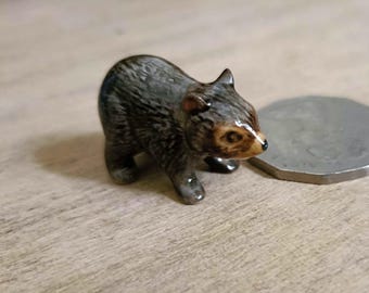 May include: A miniature ceramic bear figurine with a glossy dark brown and black finish, featuring textured fur details, small rounded ears, and a light tan muzzle with a black nose, positioned next to a silver-colored circular coin for scale.