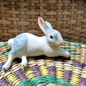 May include: A white porcelain rabbit figurine with blue accents, lying on a woven mat with green, yellow, and purple stripes.