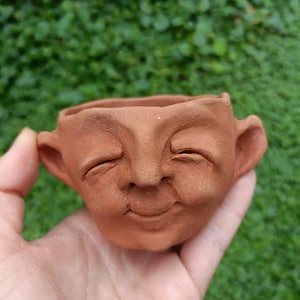 May include: A small, terracotta-colored ceramic planter shaped like a smiling face with closed eyes and large ears. The handmade pot is held in a hand against a blurred green background, suggesting an outdoor setting. The planter is designed for small plants.