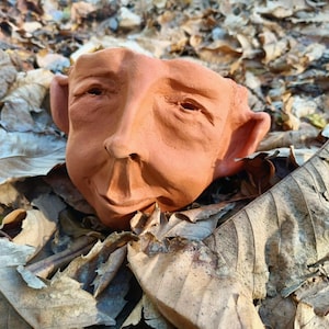 May include: A terracotta-coloured ceramic planter shaped like a human face with prominent ears, a defined nose, and closed eyes. The sculpture features a smooth, matte finish and a hollow opening at the top, showcasing detailed facial features.