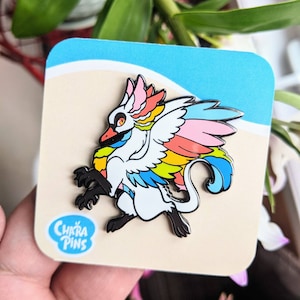 May include: A rainbow-colored enamel pin featuring a white, winged creature with a long tail. The pin is on a white card with the text "Chara Pins" in a speech bubble.