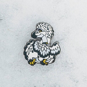 Może przedstawiać: Emaliowana szpilka w kolorze białym i czarnym przedstawiająca kreskówkowego dinozaura z żółtymi oczami i pazurami. Dinozaur jest zwinięty z złożonymi skrzydłami.