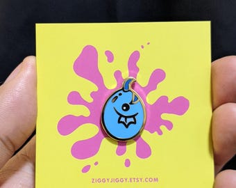 May include: A blue and gold enamel pin of a cartoon balloon with a face. The pin is on a yellow card with a pink splatter design and the text "ziggyjiggy.etsy.com".