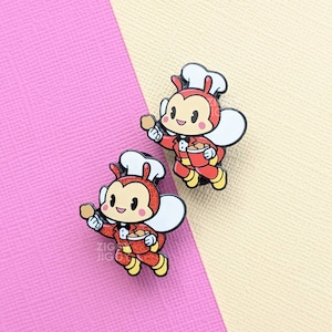 May include: Two red and black enamel pins of a cartoon bee wearing a chef's hat and holding a bucket of fried chicken. The bee is wearing a tuxedo and has a happy expression.