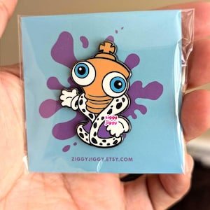 May include: A metal enamel pin featuring a cartoon character with a light orange head, large blue eyes, and a small crown, wearing a white robe with black spots and purple trim. The pin is attached to a light blue card with a purple ink splatter.